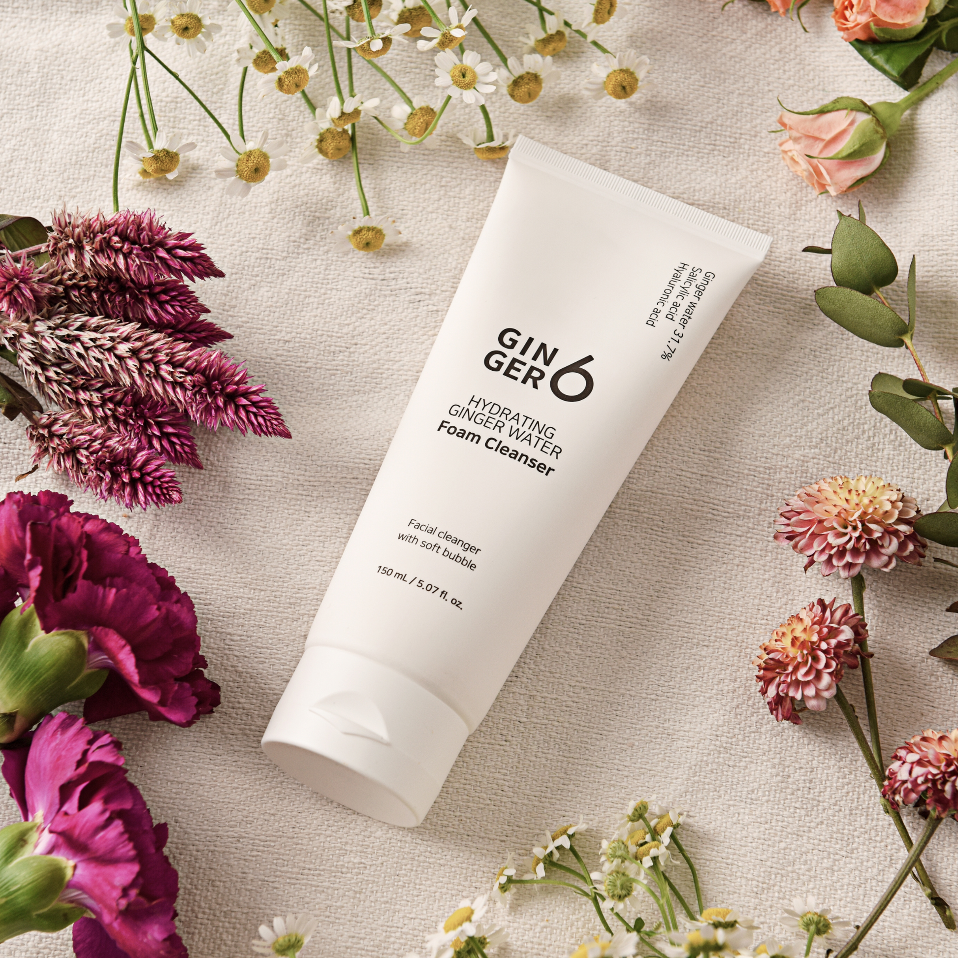 Ginger6 hydrating foam cleanser tube surrounded by fresh pink and purple flowers on a textured cloth