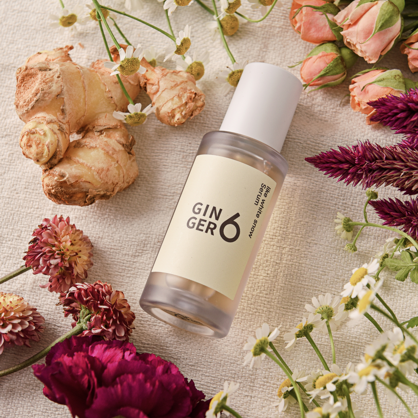 Ginger6 Like White Snow Serum surrounded by fresh pink and purple flowers on a textured cloth